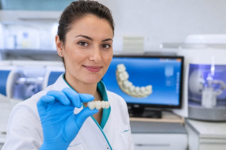 Técnica em prótese dentária em primeiro plano exibindo ponte fixa; equipamento CAD/CAM ao fundo